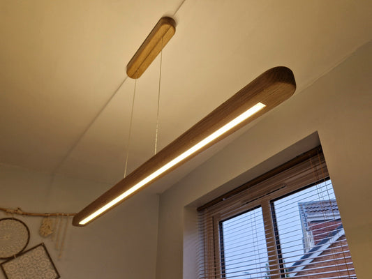 A modern linear LED pendant light with a natural wood-grain finish hangs from a thin cord in a room setting, casting a warm glow near a window with blinds.