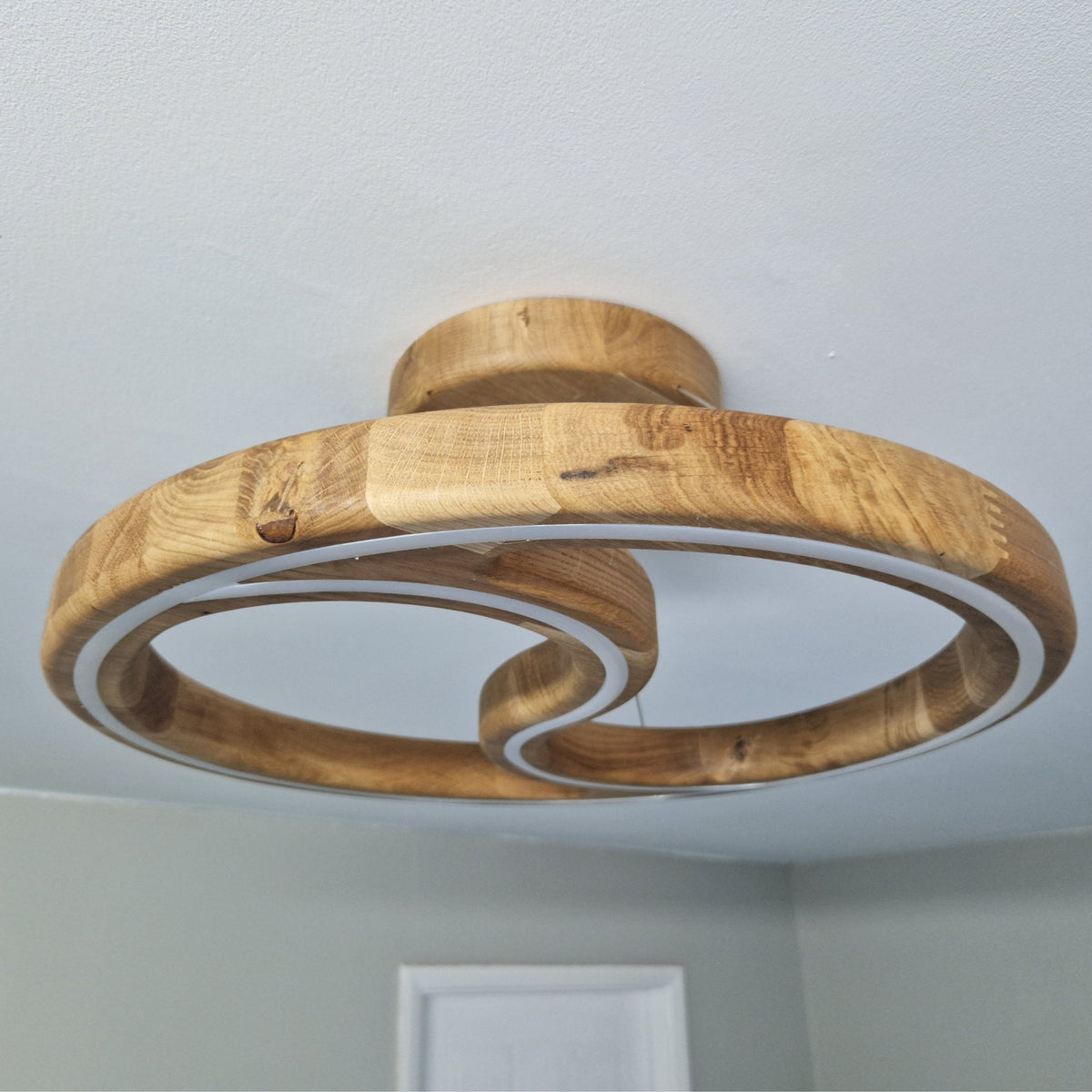 A modern spiral-shaped wooden pendant light fixture with a natural oak finish, featuring two interlocking looped rings, mounted on a white ceiling in a room.