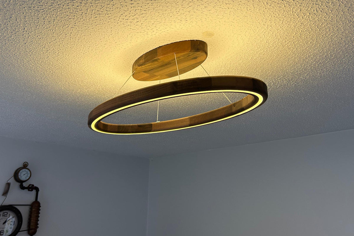 A modern double-ring LED ceiling light fixture with a warm wood finish, suspended from the ceiling in a room with a textured white ceiling and light gray walls.