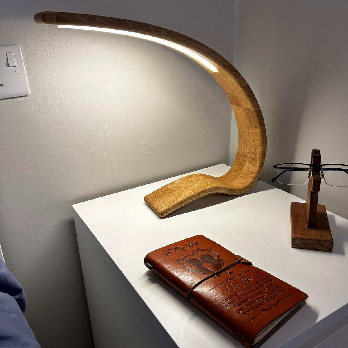 A curved wooden LED desk lamp with a warm glowing light, displayed on a white side table next to a brown leather journal and a wooden eyeglasses stand.