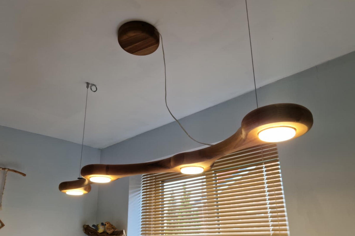 A modern multi-light linear pendant chandelier with a curved wooden body and four glowing round bulbs, suspended from the ceiling in a room with light blue walls and window blinds.