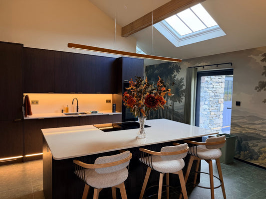 Modern kitchen with a bespoke wooden linear pendant light above a large island, warm layered lighting, dark wood cabinetry, skylight, natural stone finishes and contemporary dining stools.