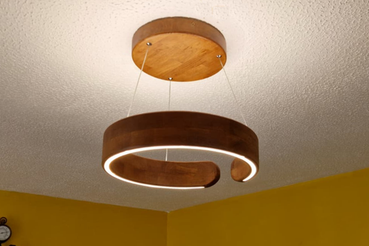 A modern two-tier wooden ring pendant light with an illuminated white LED strip, suspended from a round wooden ceiling mount against a textured white ceiling and yellow wall.