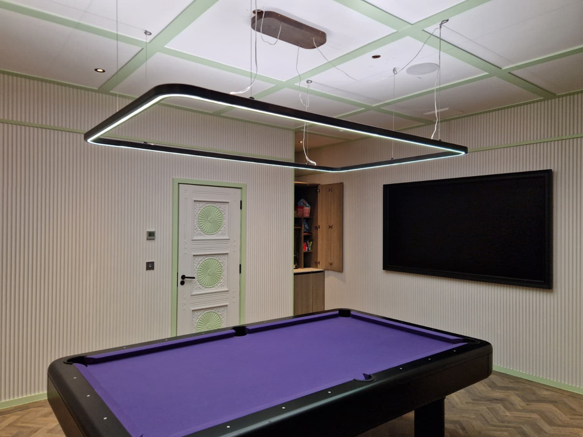 A modern rectangular LED undercabinet light with a black frame and white light strip, suspended from the ceiling of a room with a purple pool table.