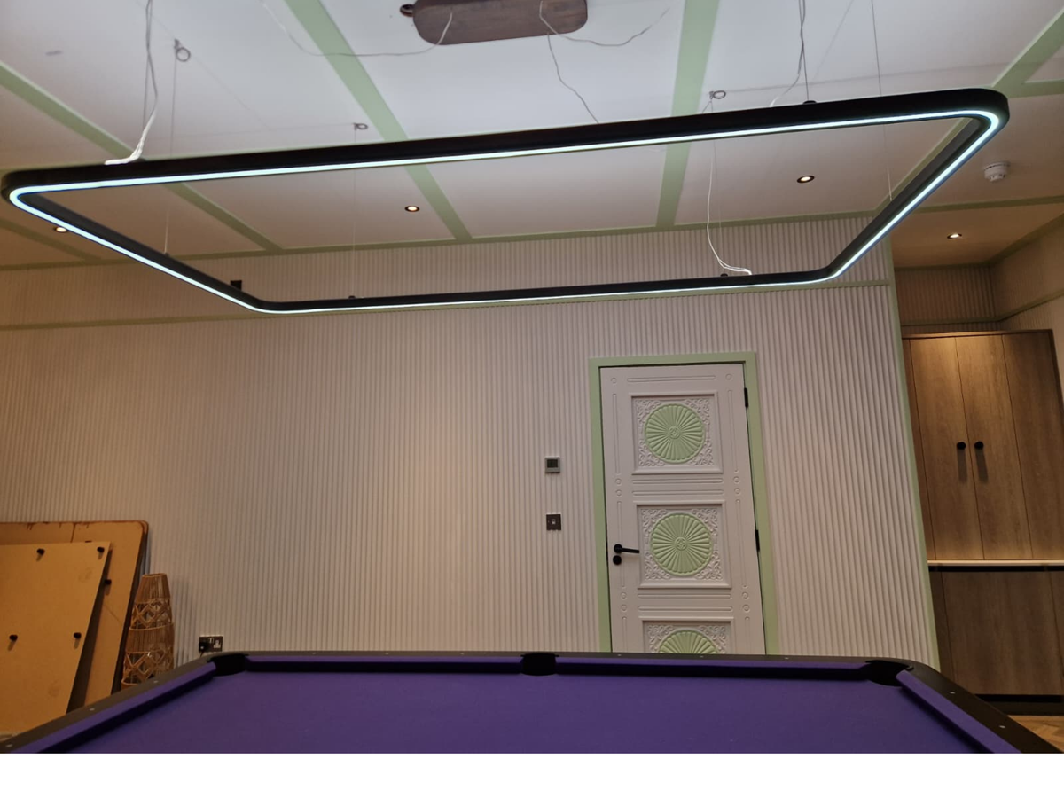 A large rectangular LED undercabinet light with a black frame and glowing white light bars, suspended from the ceiling of a modern room with a purple pool table in the foreground.