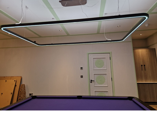 A large rectangular LED undercabinet light with a black frame and glowing white light bars, suspended from the ceiling of a modern room with a purple pool table in the foreground.