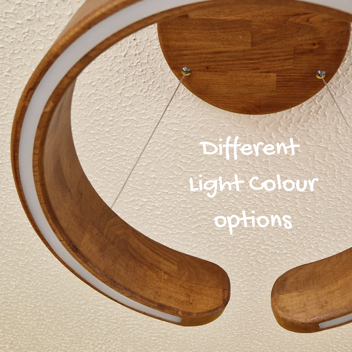 Close-up of a modern curved wooden ceiling light fixture with a white inner ring, mounted against a textured cream wall. White text in the center reads 'Different Light Colour options'.