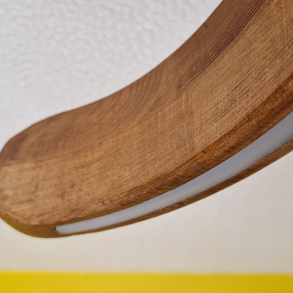 Extreme close-up of the curved oak wood pendant light showing the wood grain and the white illuminated LED strip along the underside, set against an off-white textured wall with a yellow surface visible at the bottom.