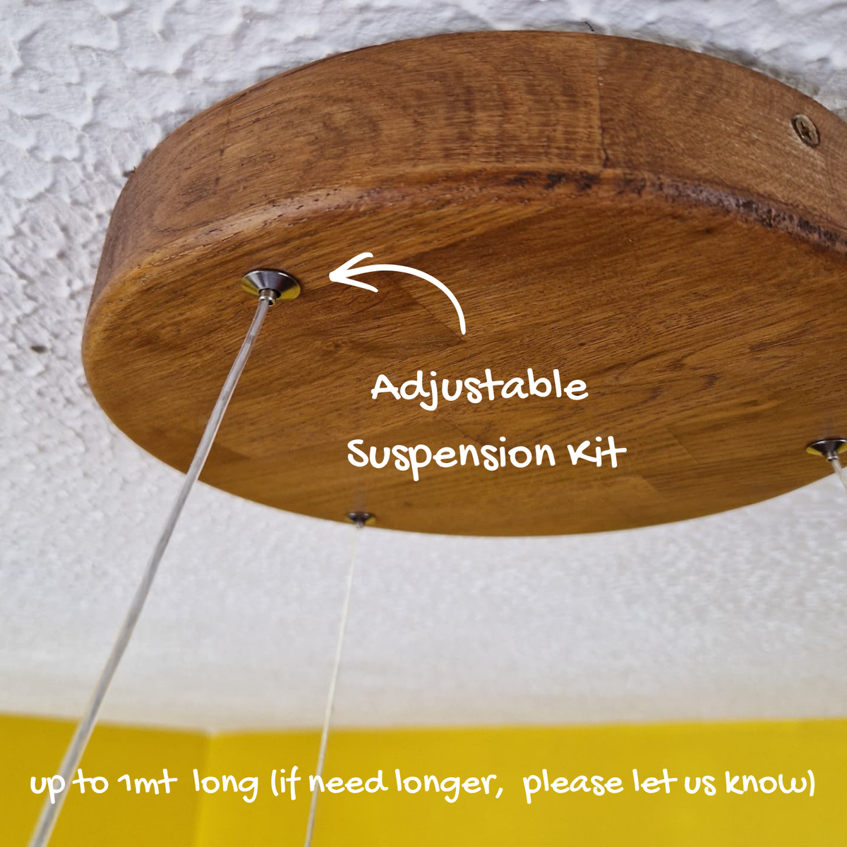 A close-up of a round wooden ceiling mount with hanging suspension wires and overlaid text reading 'Adjustable Suspension Kit' with a note about cord length, shown against a textured white ceiling and yellow wall.