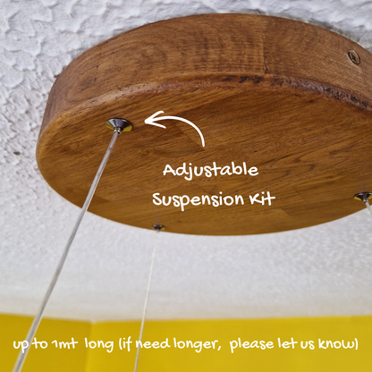A close-up of a round wooden ceiling mount with hanging suspension wires and overlaid text reading 'Adjustable Suspension Kit' with a note about cord length, shown against a textured white ceiling and yellow wall.