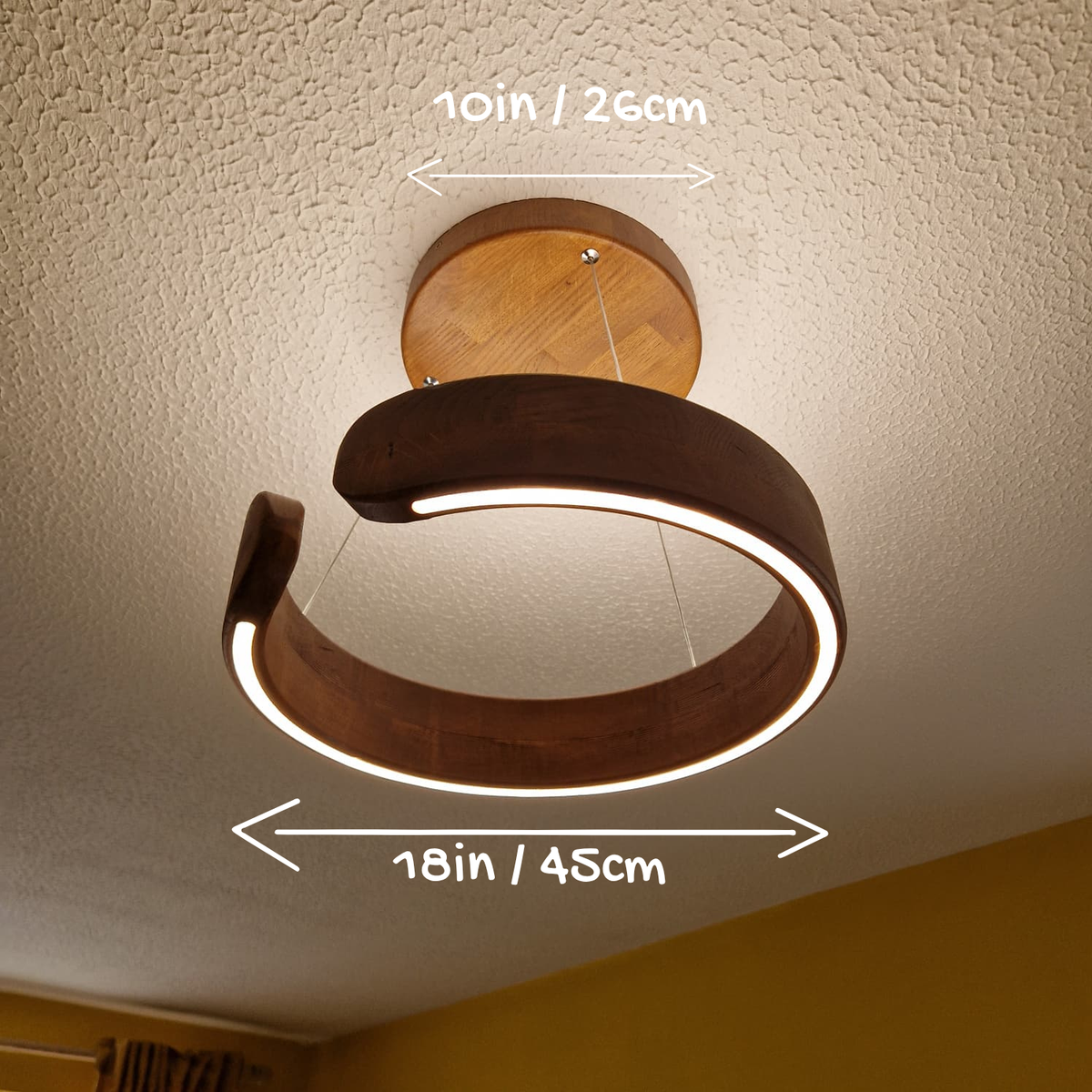 A modern spiral-shaped LED ceiling light fixture with a dark wood frame and circular wood ceiling mount, shown installed on a textured white ceiling with dimension arrows indicating 18 inches wide by 10 inches deep.