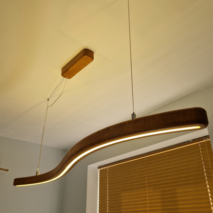 A modern curved LED linear pendant light with a dark wood-grain finish, suspended from the ceiling in a room with window blinds.