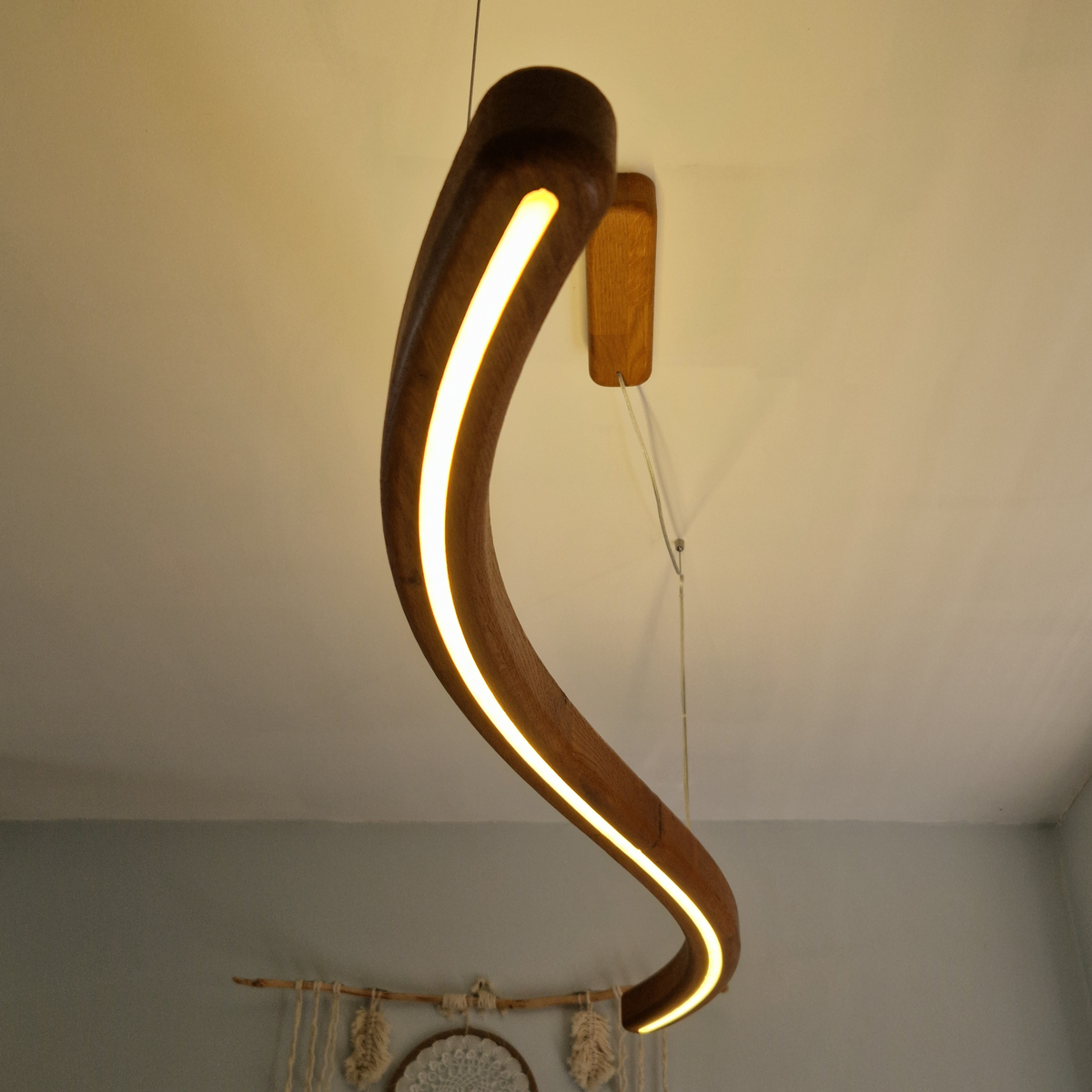 A curved wooden pendant light with a warm glowing LED strip, suspended from a ceiling in an indoor room setting.