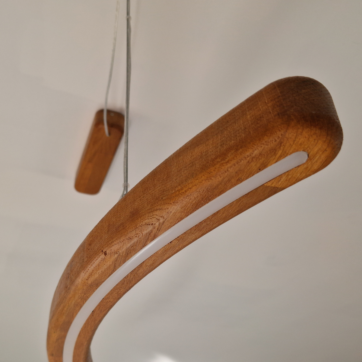 A curved wooden pendant light fixture with a warm medium-brown wood grain finish and a white frosted light strip, shown hanging against a plain off-white wall.