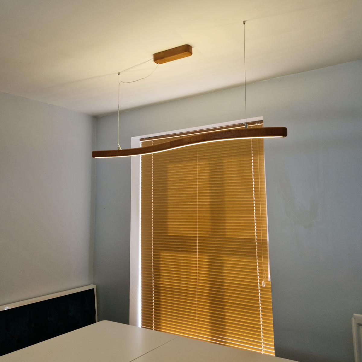 A modern linear pendant light with a curved wooden frame and integrated LED strip, suspended from the ceiling in a room with light blue-gray walls and a window with closed blinds.