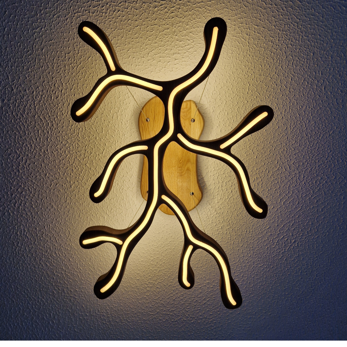 A modern wall sconce light fixture with a branching, tree-like silhouette made of black material, glowing warm yellow LED lines, and a natural wood backplate, shown against a dark textured wall.