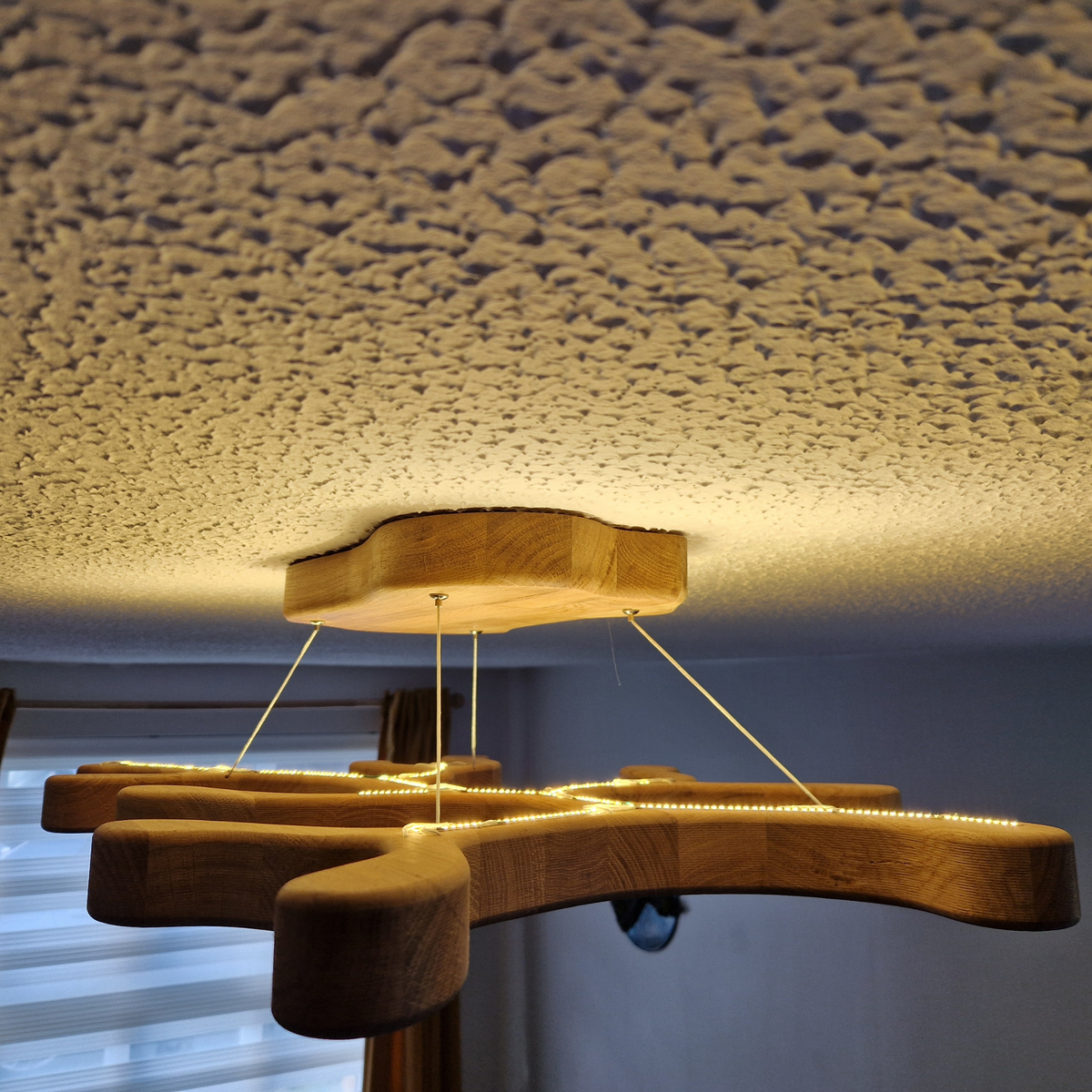 A modern wooden ceiling light fixture with a geometric, branch-like silhouette and warm LED strip lighting, suspended from a square wooden ceiling plate in a home interior.