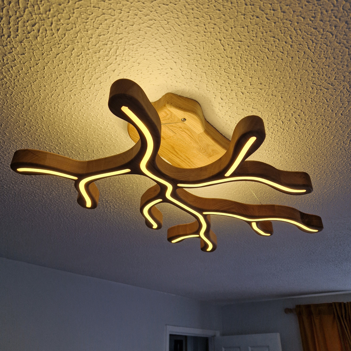 A modern branch-shaped ceiling light fixture made of natural wood with integrated warm white LED lighting, shown installed on a textured white ceiling in a room.