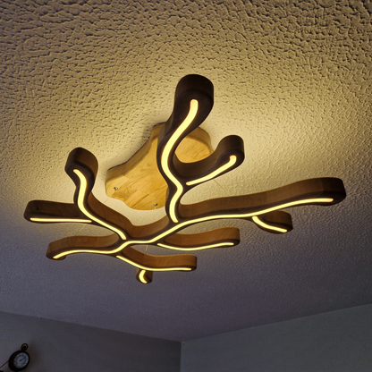 A modern LED light fixture with organic, branching coral-like wooden arms emitting warm yellow light, mounted on a textured white ceiling.