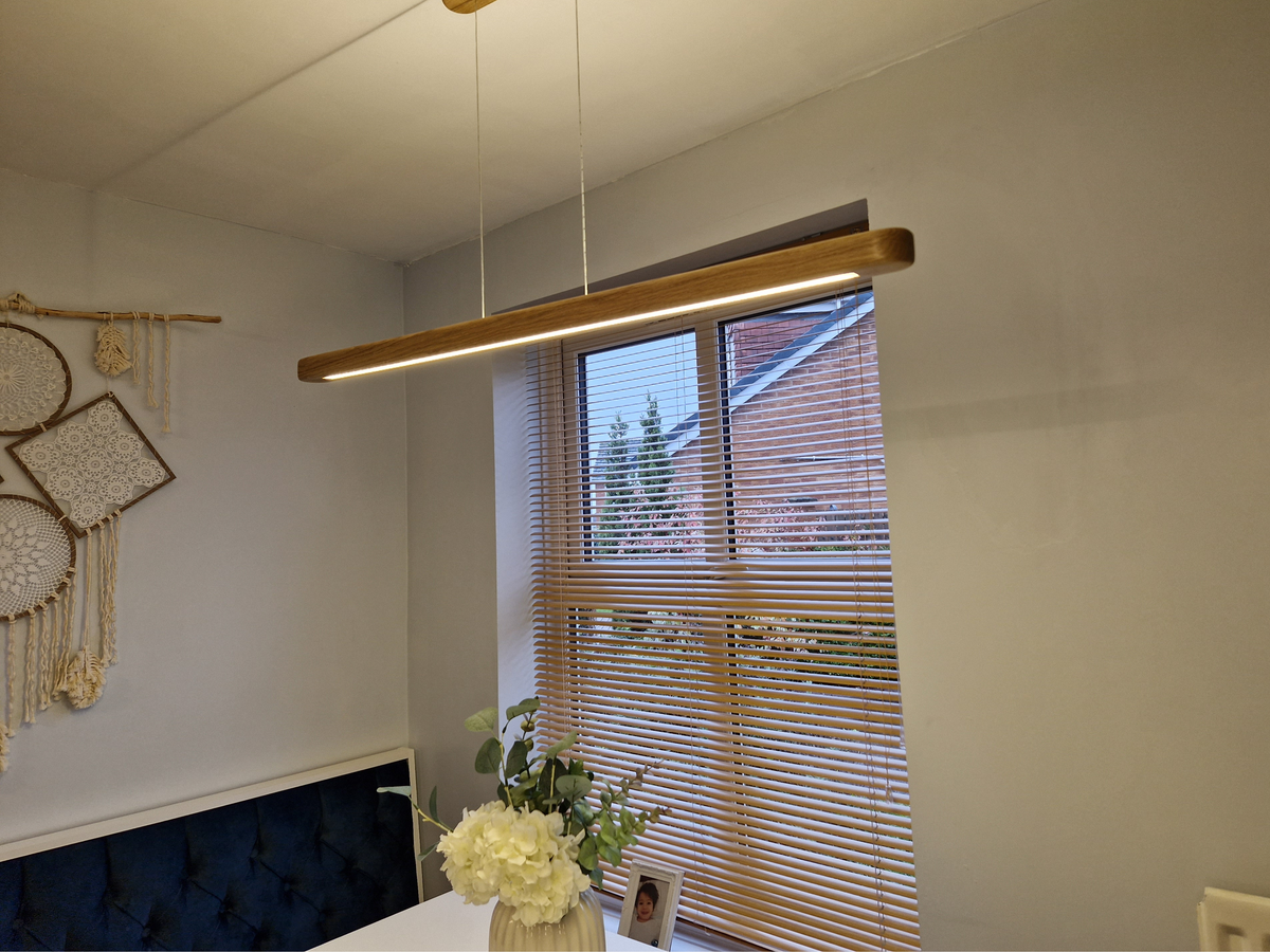 A modern linear pendant light with a natural oak wood finish and integrated LED strip, hanging in a dining or living room setting next to a window with wooden blinds.