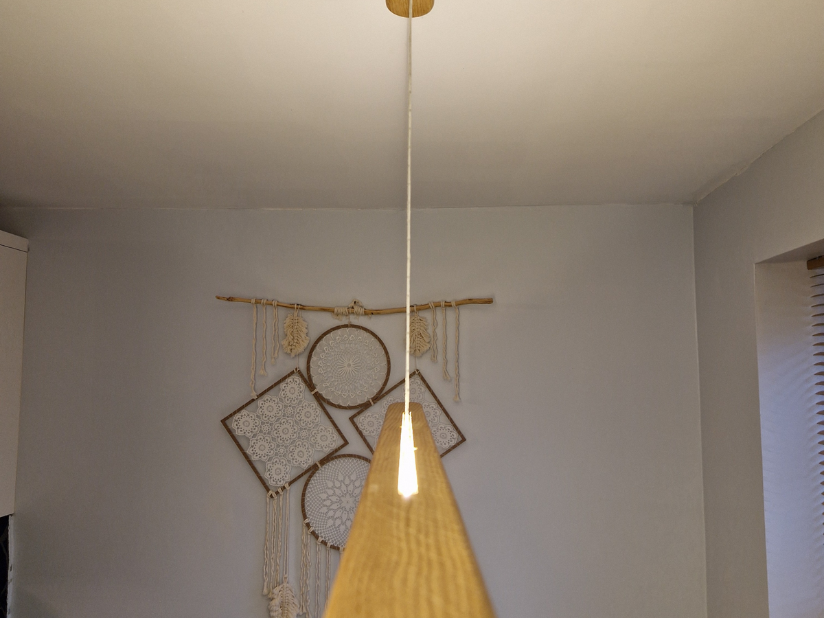 A modern pendant light with a tapered wooden shade and a single white cord hanging from a gold ceiling mount, set against a neutral wall with boho dreamcatcher decor.