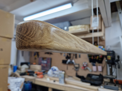 A close-up of a curved, tapered solid oak wood pendant light component hanging in a woodworking workshop, surrounded by tools and equipment.