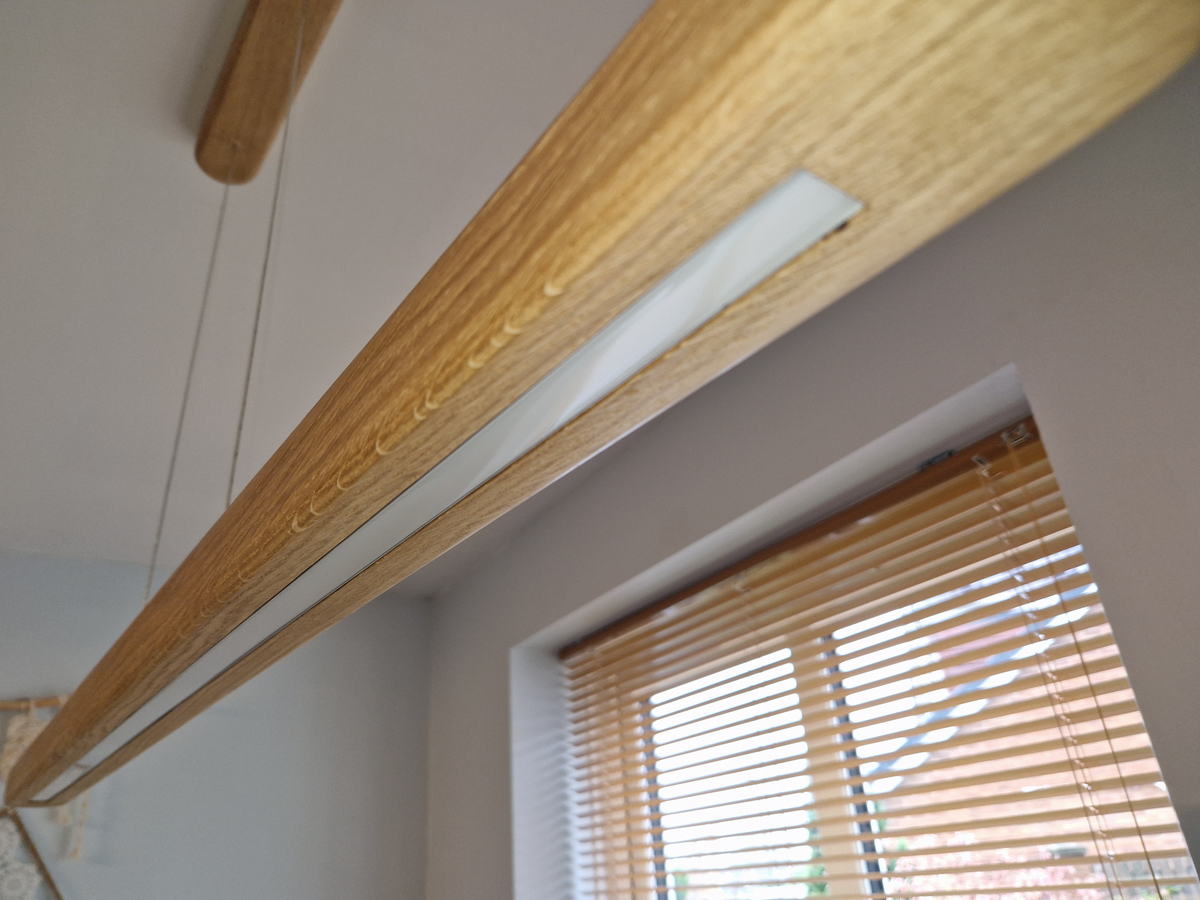 A close-up of a modern linear LED light fixture with a natural oak wood frame and a white diffuser, shown installed in a ceiling near a window with wooden blinds.