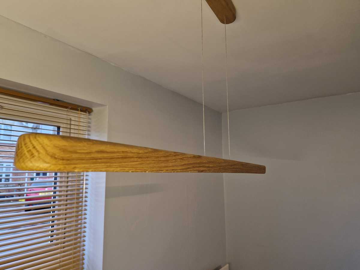 A modern minimalist linear pendant light made of natural oak wood, suspended by two thin white cables from a matching ceiling mount, shown in a minimalist room setting.