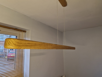A modern minimalist linear pendant light made of natural oak wood, suspended by two thin white cables from a matching ceiling mount, shown in a minimalist room setting.
