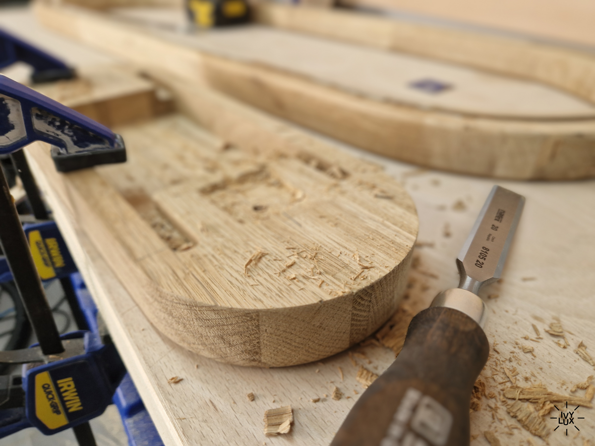 A close-up of a wooden chisel resting on a workbench surrounded by wood shavings, with a blue and black clamp holding a curved wooden piece in the foreground and more wood pieces blurred in the background.