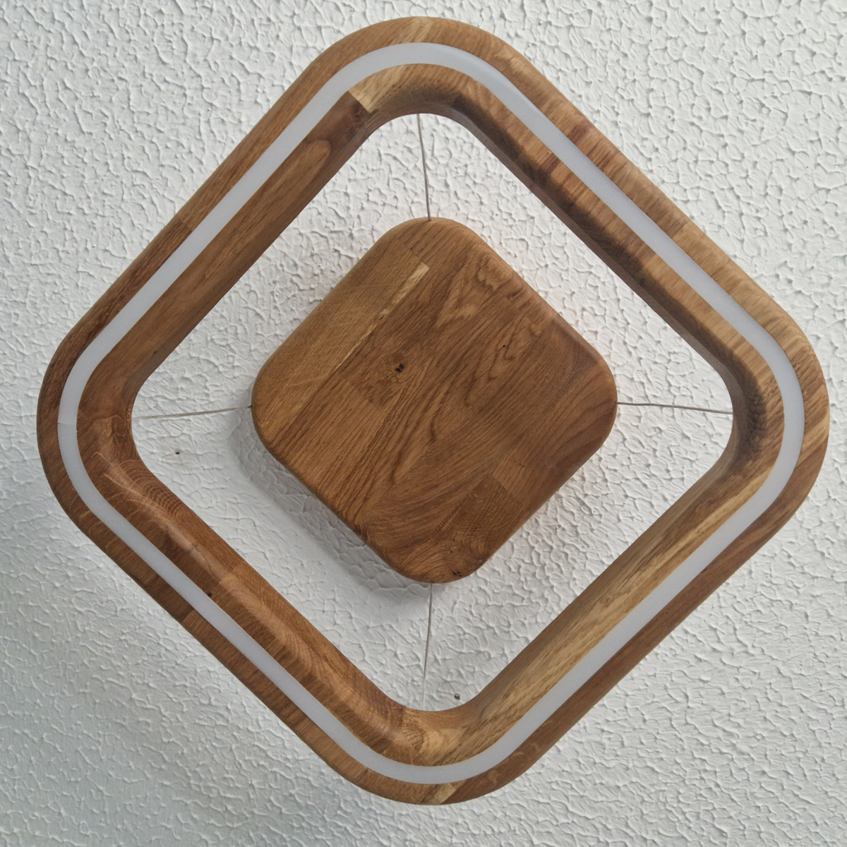 A modern geometric ceiling-mounted light fixture with a square wooden frame in a diamond orientation, featuring a central square wooden panel and a white LED light strip along the inner border, shown against a textured white ceiling, viewed from directly below.