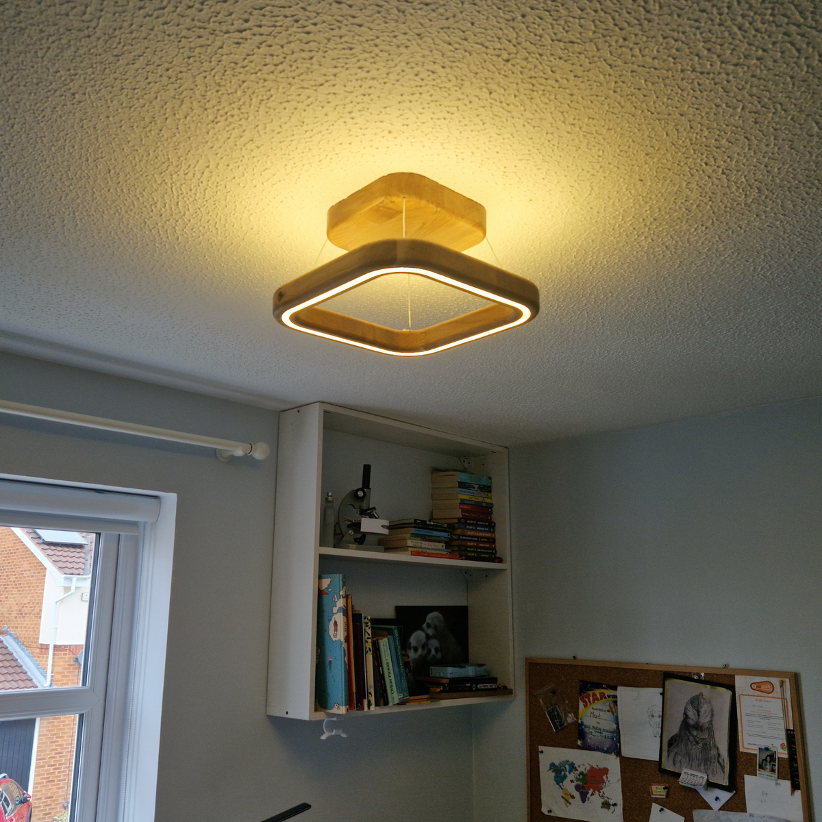 A modern square LED ceiling light with a warm yellow glow, mounted on a textured white ceiling in a room with a built-in white shelf and a window.