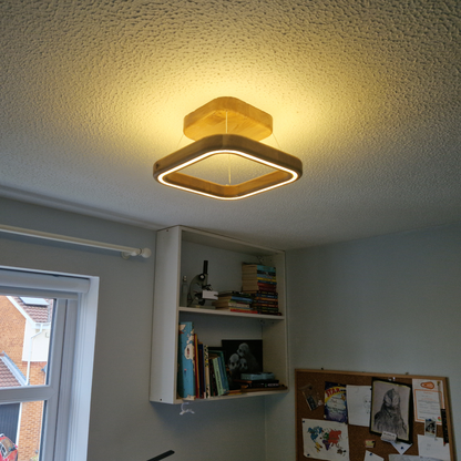 A modern square LED ceiling light with a warm yellow glow, mounted on a textured white ceiling in a room with a built-in white shelf and a window.