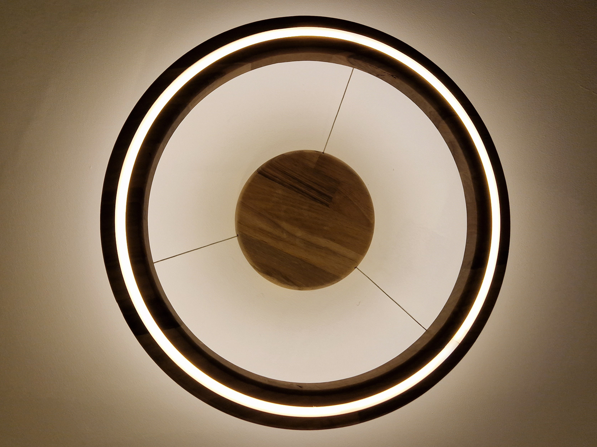 A modern circular ceiling light fixture with a glowing warm white LED ring and a natural wood central hub, photographed from directly below against a matching light-colored ceiling.