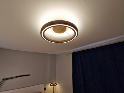 A modern circular ceiling light fixture with a warm wood finish and a glowing white LED ring, installed on a white ceiling in a room with a dark blue curtain.