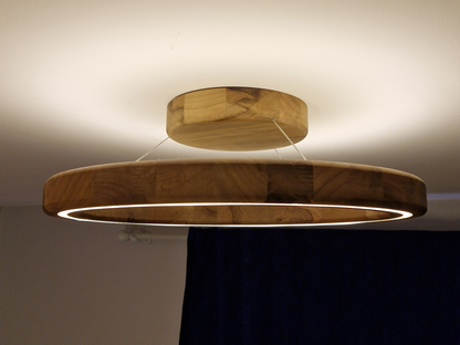 A modern circular wooden ceiling light fixture with a warm LED ring, suspended by thin cables from a matching round ceiling mount, shown against a white ceiling and dark blue curtain.