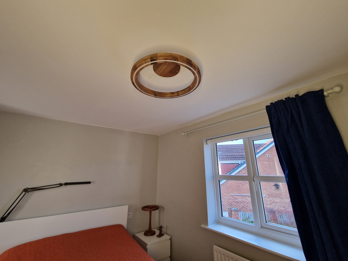 A modern circular ceiling light fixture with a warm wood-grain outer ring and a white inner ring, mounted on a white ceiling in a bedroom setting.
