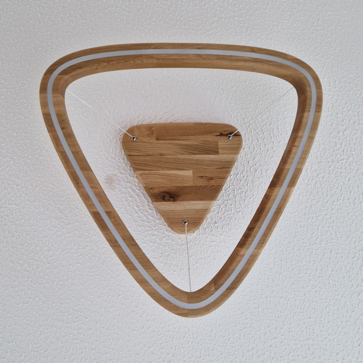 A triangular wooden LED pendant light viewed from directly below, showing an open triangle ring frame around a smaller triangular wooden ceiling mount, installed on a textured white ceiling.