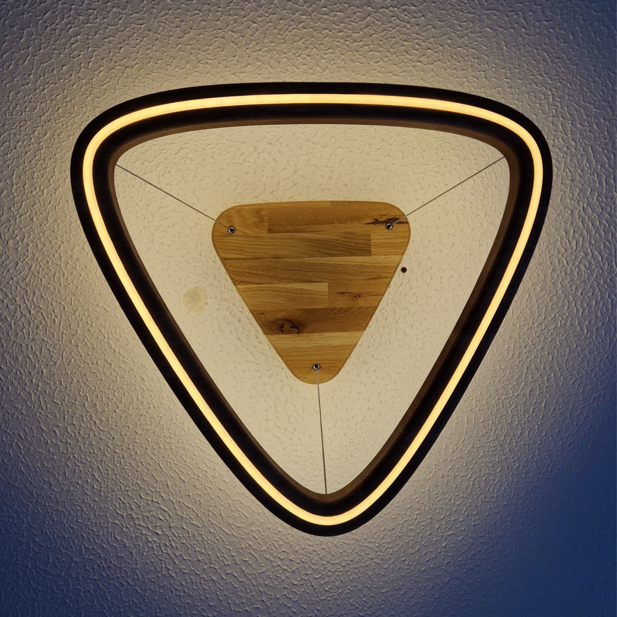 Triangular LED pendant light fixture viewed from directly below, with a glowing warm white LED outer ring, a black frame, and a natural wood central panel, suspended from a textured ceiling.