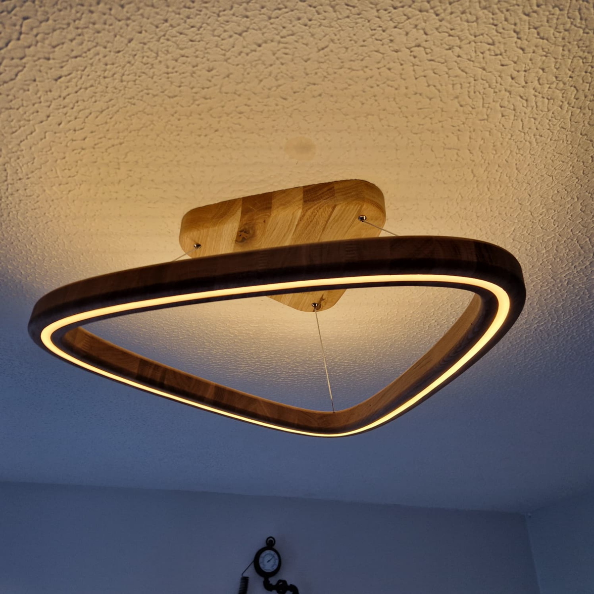 A modern triangular LED ceiling light with a dark wood frame and a warm glowing light, mounted on a textured white ceiling.