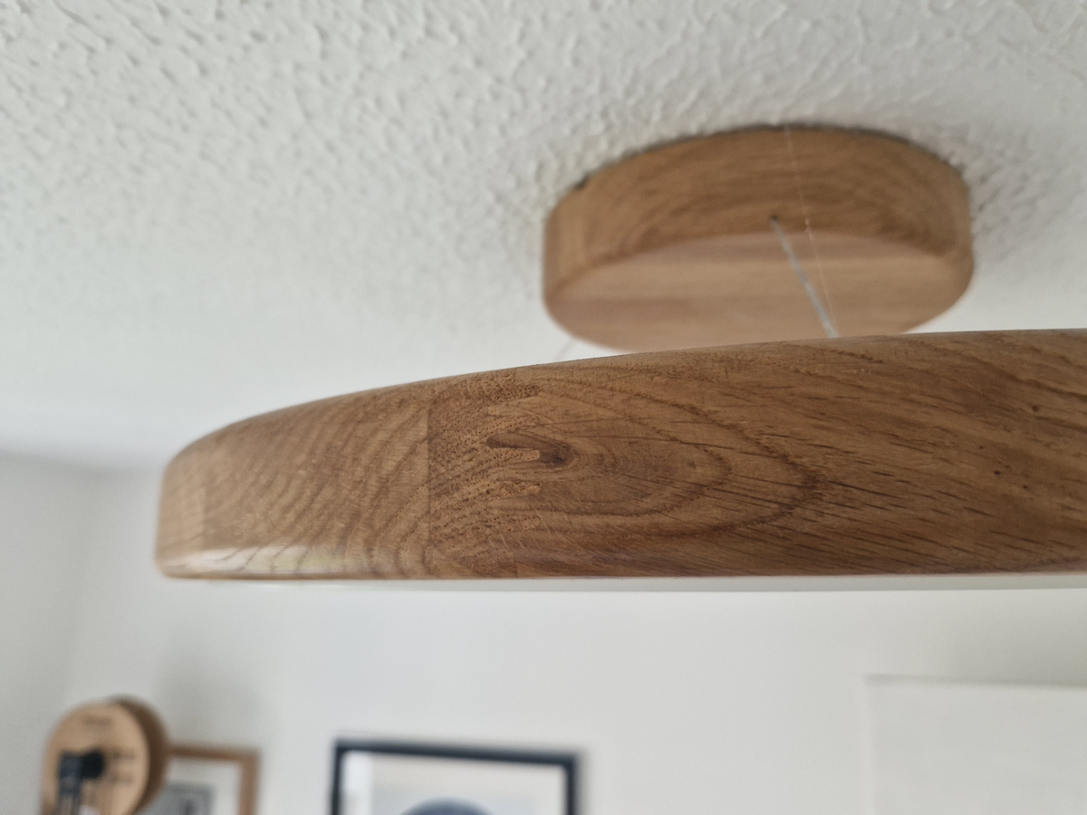 A close-up of a round wooden ceiling light fixture with a visible wood grain, suspended by a thin cord from a circular ceiling mount against a white textured ceiling.