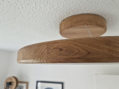 A close-up of a round wooden ceiling light fixture with a visible wood grain, suspended by a thin cord from a circular ceiling mount against a white textured ceiling.