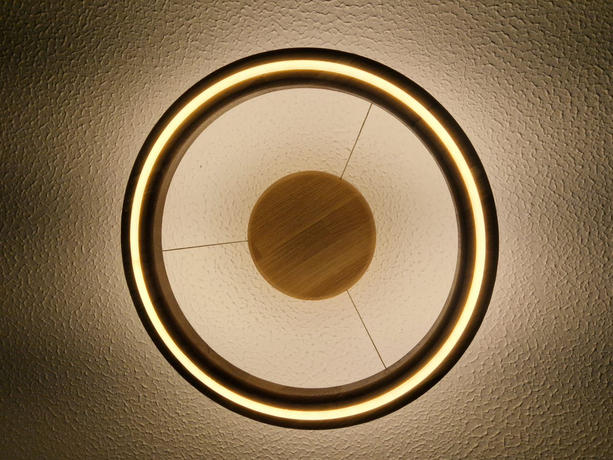 A modern circular ceiling light fixture with a glowing warm white LED ring, a textured white inner panel, and a central round wooden accent, shown mounted on a textured beige ceiling, viewed from directly below.
