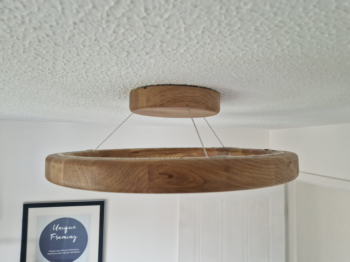 A circular wooden pendant light with a thick oak-look finish hangs from thin cables on a textured white ceiling in a home interior setting.