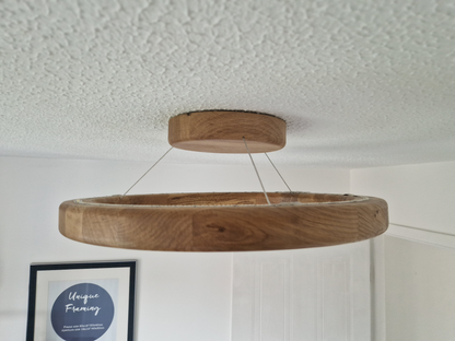 A circular wooden pendant light with a thick oak-look finish hangs from thin cables on a textured white ceiling in a home interior setting.