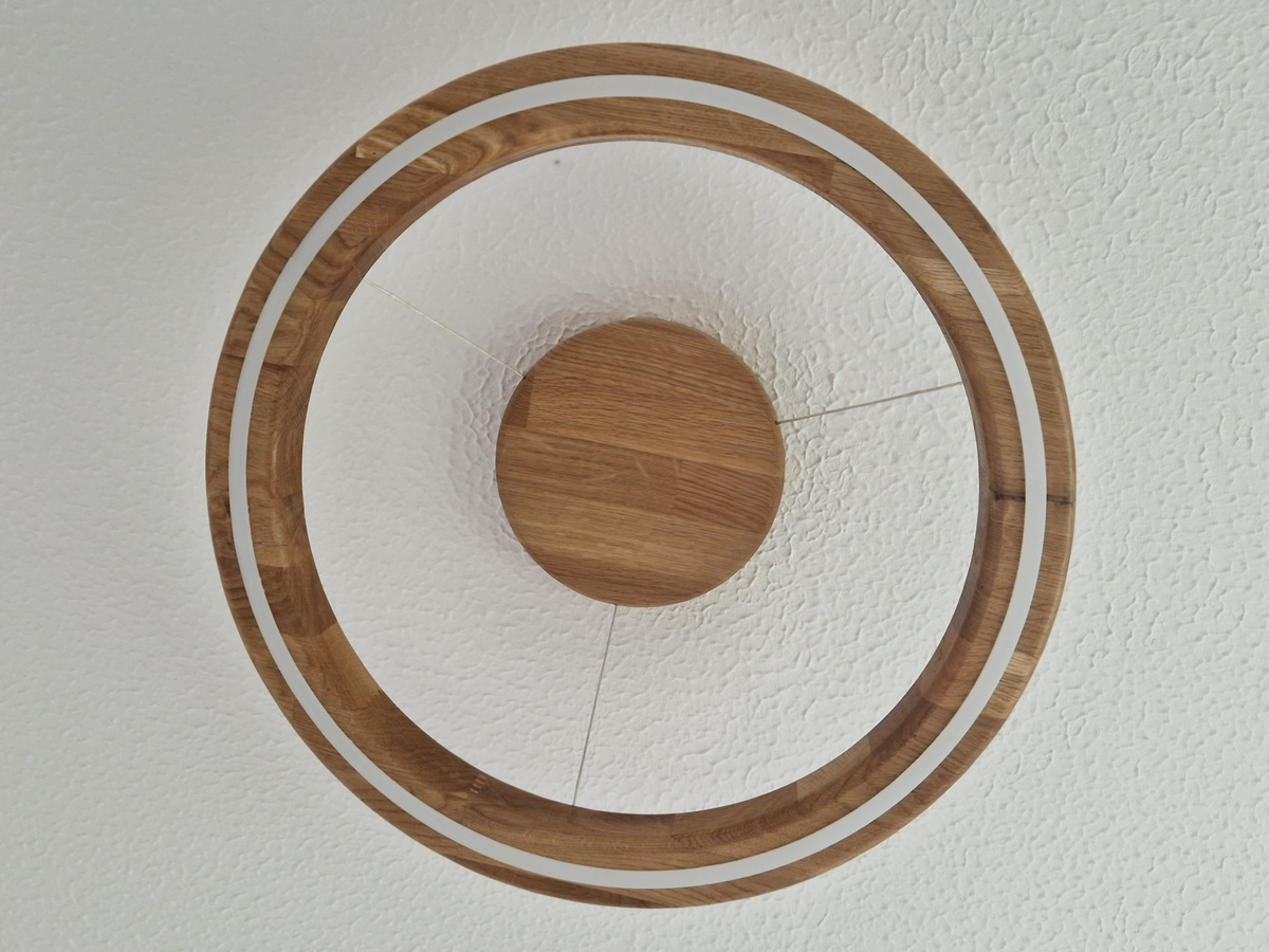 A circular wooden ceiling light fixture viewed from directly below, featuring a wood outer ring with white LED accent trim and a round wood center mount, suspended by thin cables against a textured white ceiling.