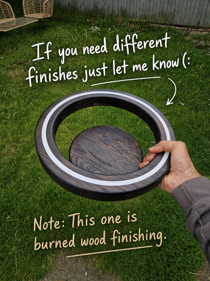 A hand holds a round wooden tray or coaster outdoors on grass, with overlaid text saying 'If you need different finishes just let me know (:' and 'Note: This one is burned wood finishing.'.