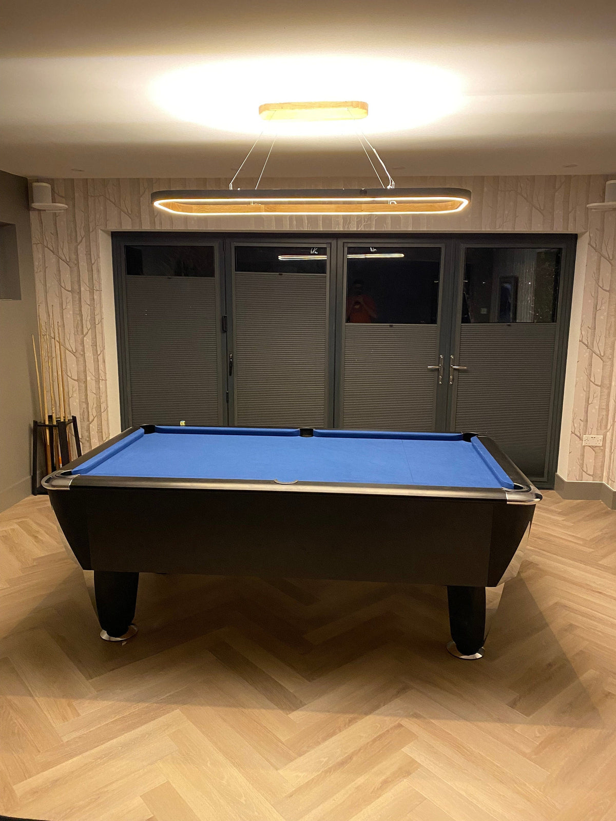 A pool table with a blue felt surface and dark frame, placed in a room with light wood herringbone flooring, paneled walls, and a large rectangular ceiling light above.
