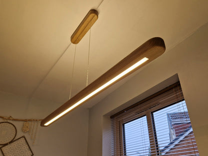 A modern linear LED pendant light with a natural wood-grain finish hangs from a thin cord in a room setting, casting a warm glow near a window with blinds.