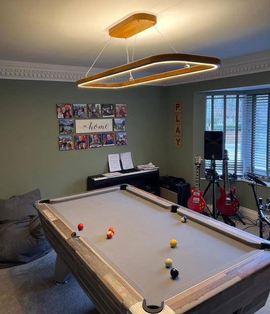 A modern rectangular LED pendant light with a warm wood frame hangs over a pool table in a home entertainment room with guitars, a piano, and a beanbag chair.